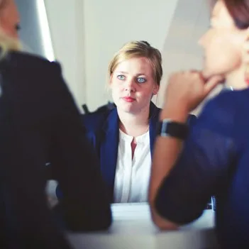 A woman sitting slightly back from a meeting table, listening to her colleagues