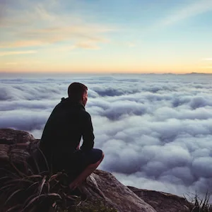 A man sitting on a rock, facing a sea of clouds in the morning light