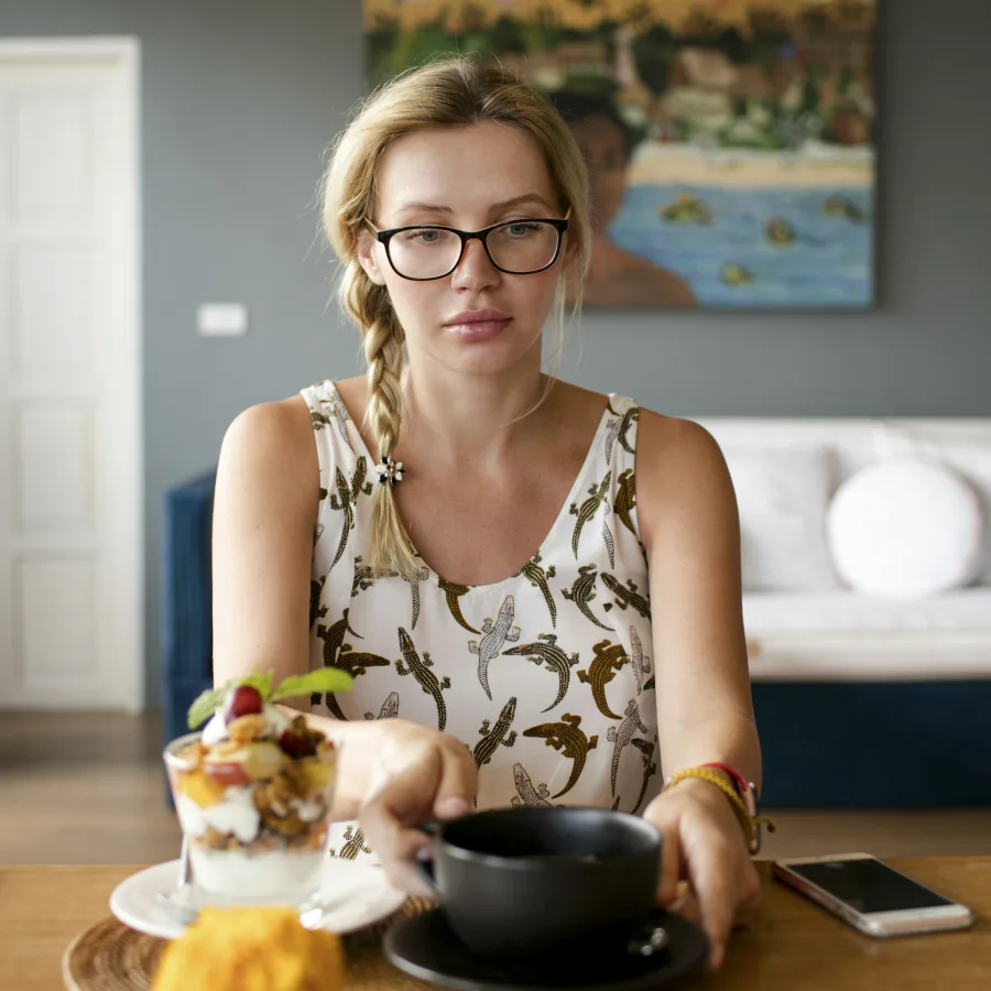 A young woman in glasses, looking worried, sitting at a table with a coffee and a fruit cup