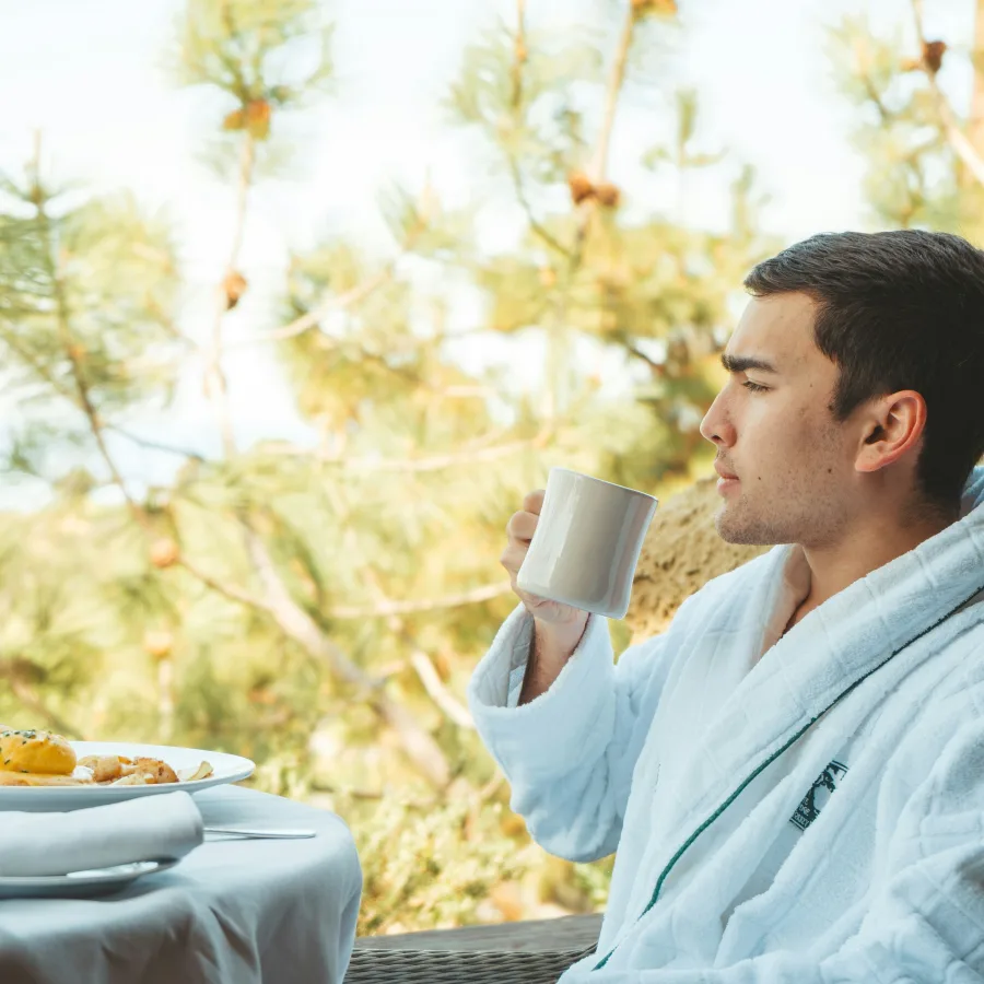 Joven en albornoz tomando su café de la mañana en una terraza, frente a los pinos