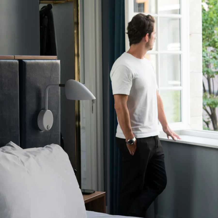 Hombre con camiseta blanca, de pie junto a la ventana de un dormitorio, mirando hacia fuera