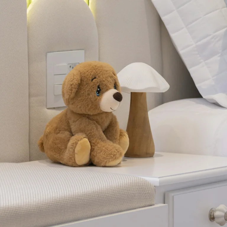 A teddy bear sitting at the edge of a child's bed, beside a small lit lamp