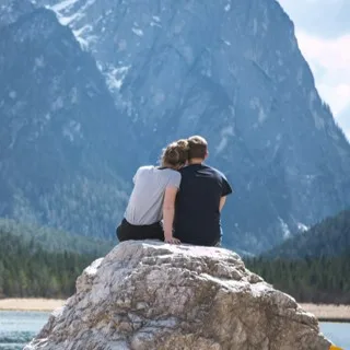 Two people sitting side by side on a rock, head against shoulder, facing the mountains
