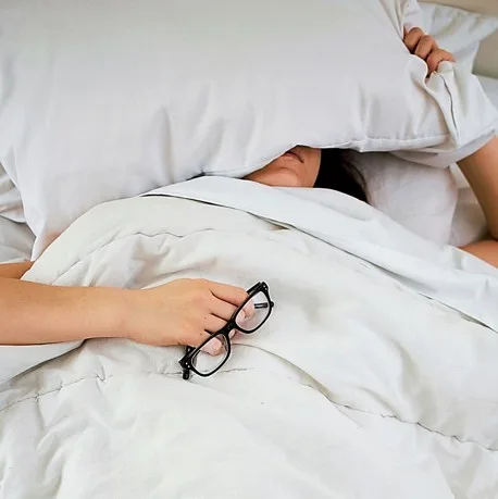 Person lying under a pillow, glasses in hand