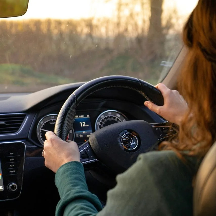 Las manos de una conductora apoyadas en el volante, en una carretera rural al atardecer