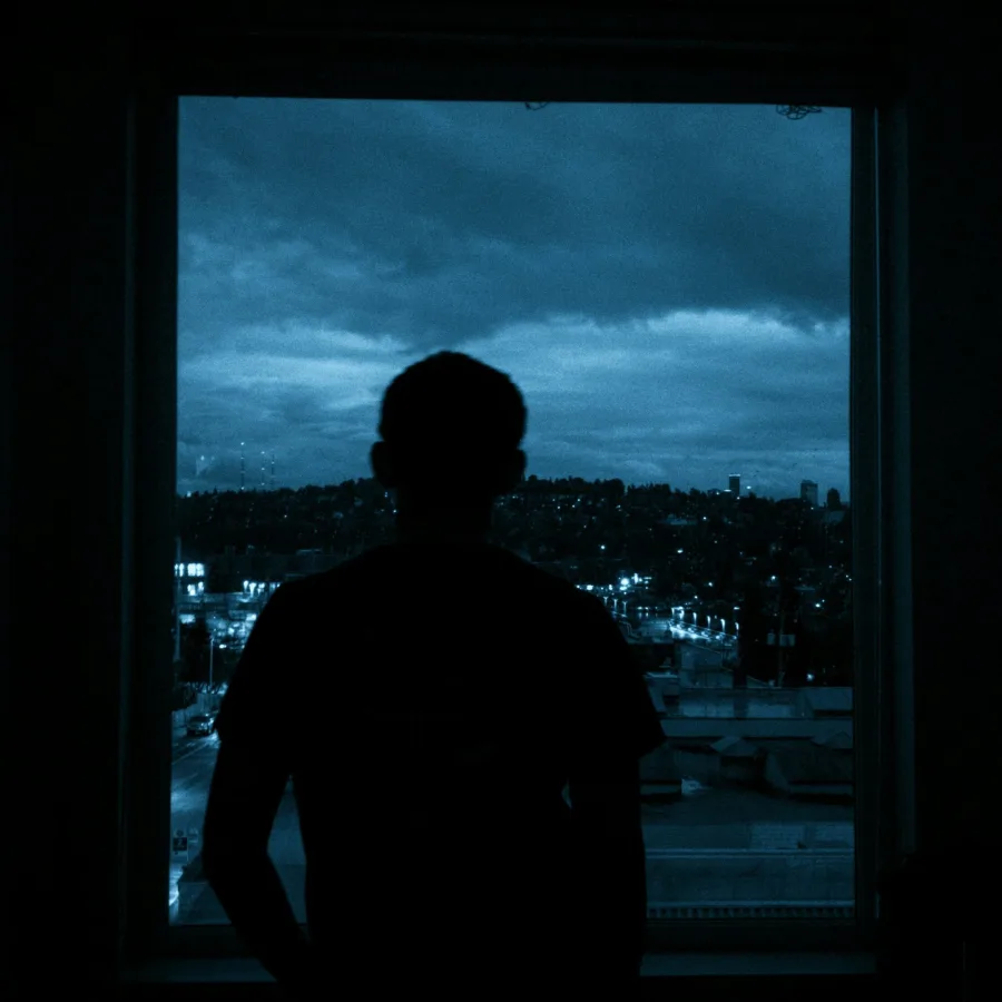 Silueta sola ante una ventana de noche, interior oscuro, luces de la ciudad a lo lejos