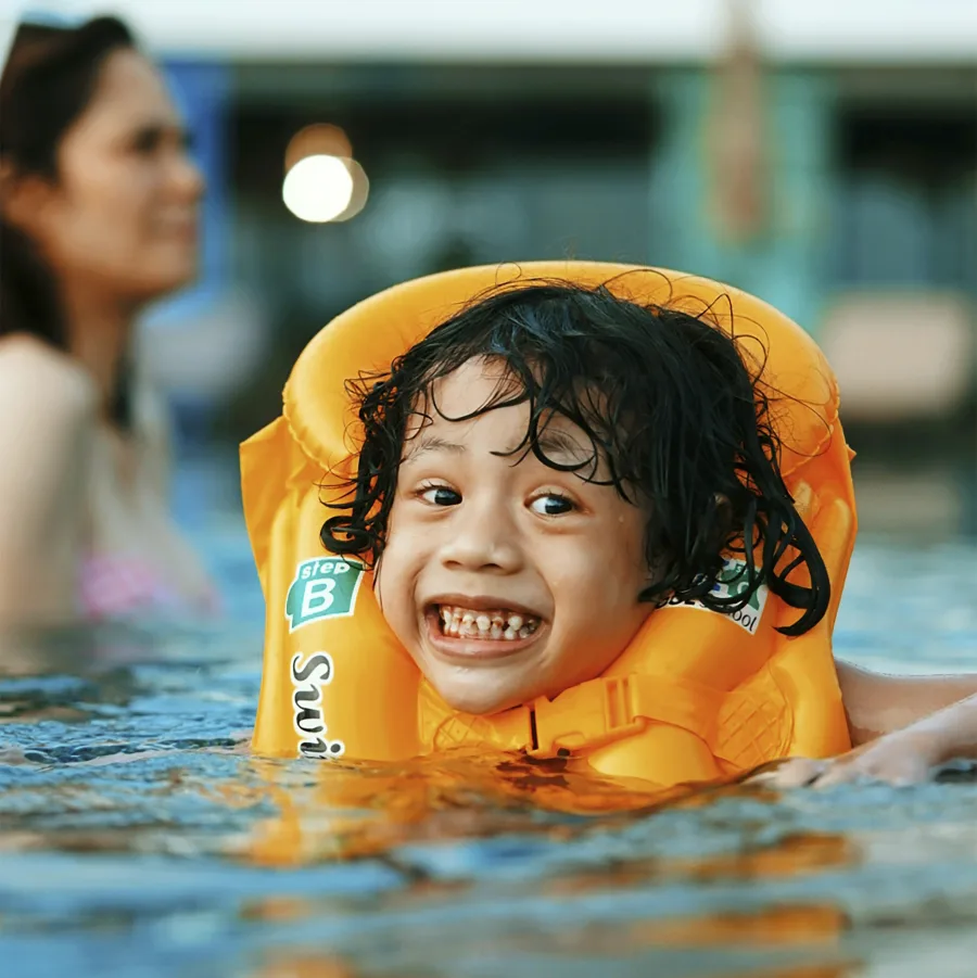 Smiling child in a swimming pool wearing a flotation vest, an adult nearby
