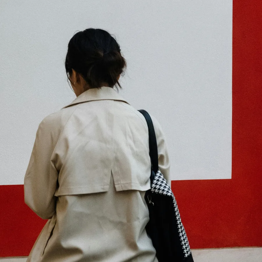 Mujer de espaldas, con abrigo claro y bolso al hombro, ante una pared blanca y roja