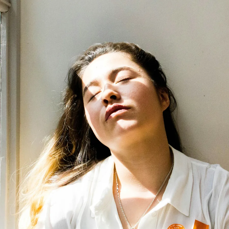 A woman leaning against a wall near a window, eyes closed, her face in the light