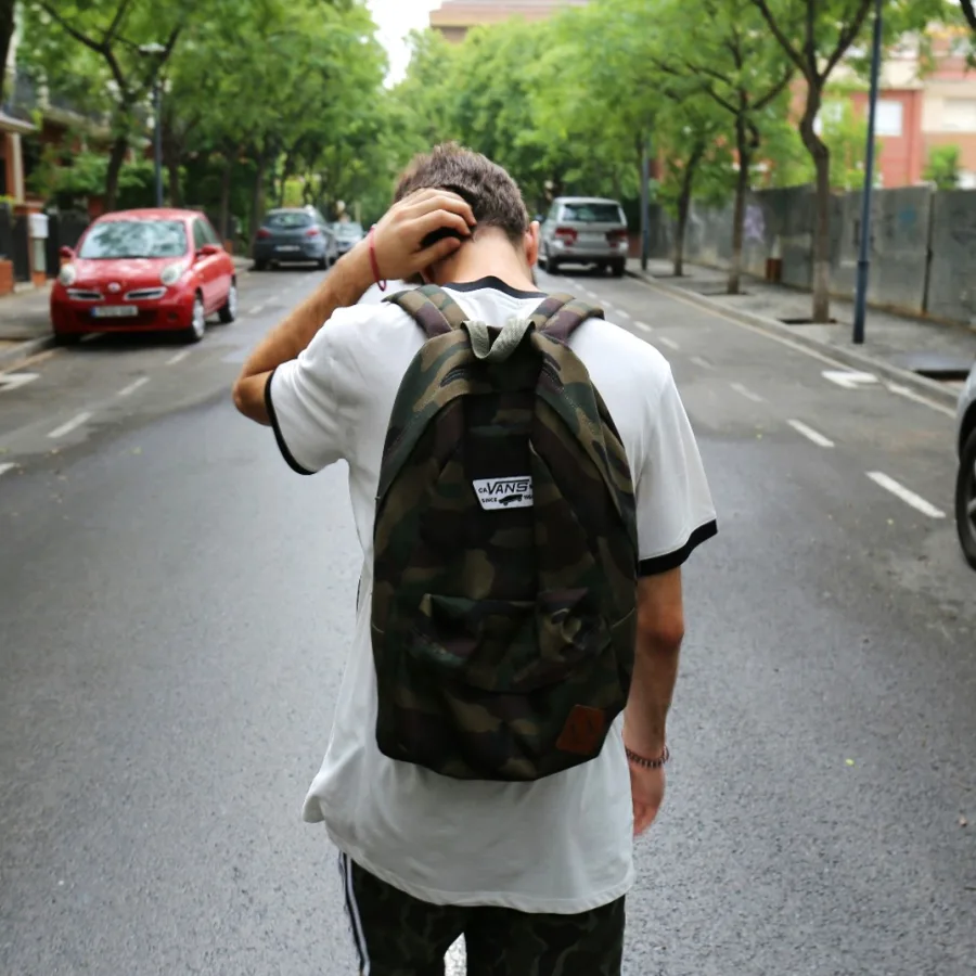 A teenager seen from behind in the street, backpack on, one hand on the back of his neck