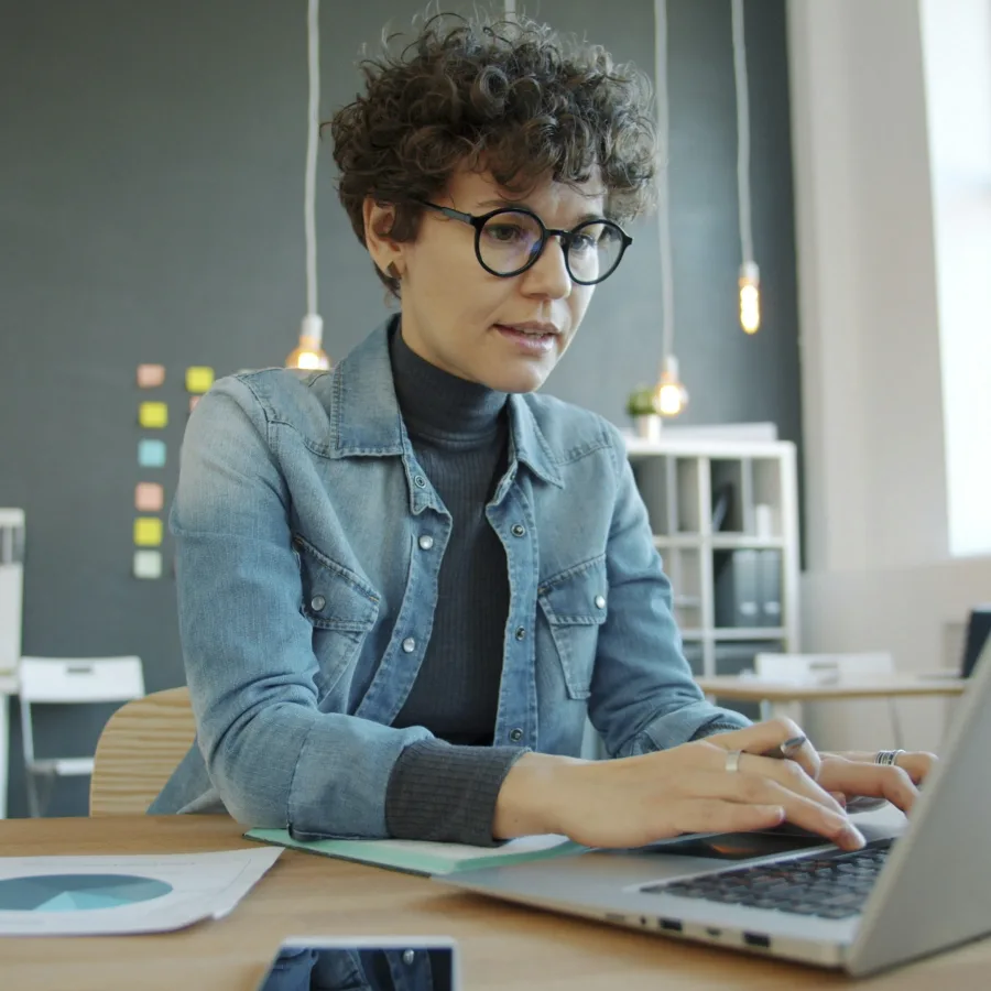 A woman with curly hair and round glasses, absorbed in her laptop in a bright office