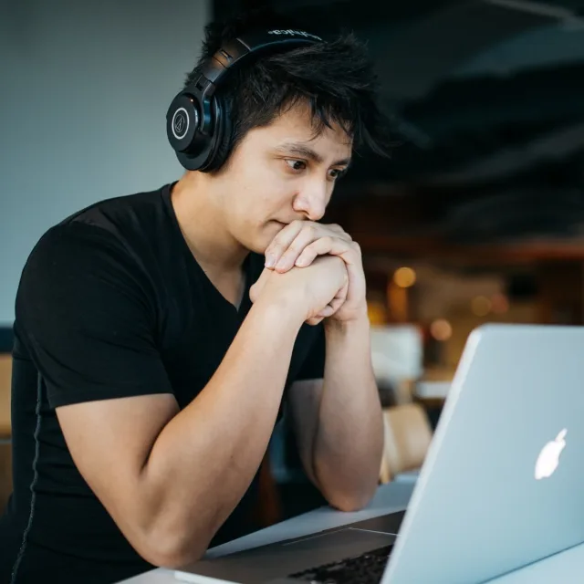 Young man with headphones on, forehead resting on his hands, staring tensely at his laptop