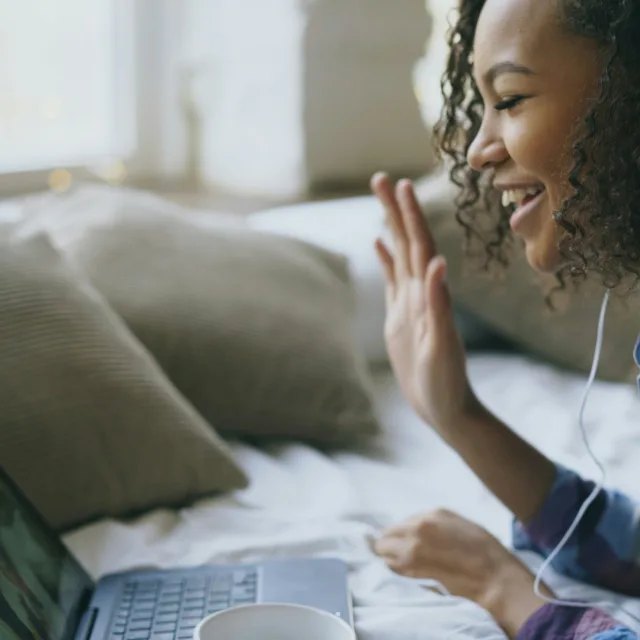 A young woman lying on her bed, wearing earphones, waving at the person she can see on her laptop screen
